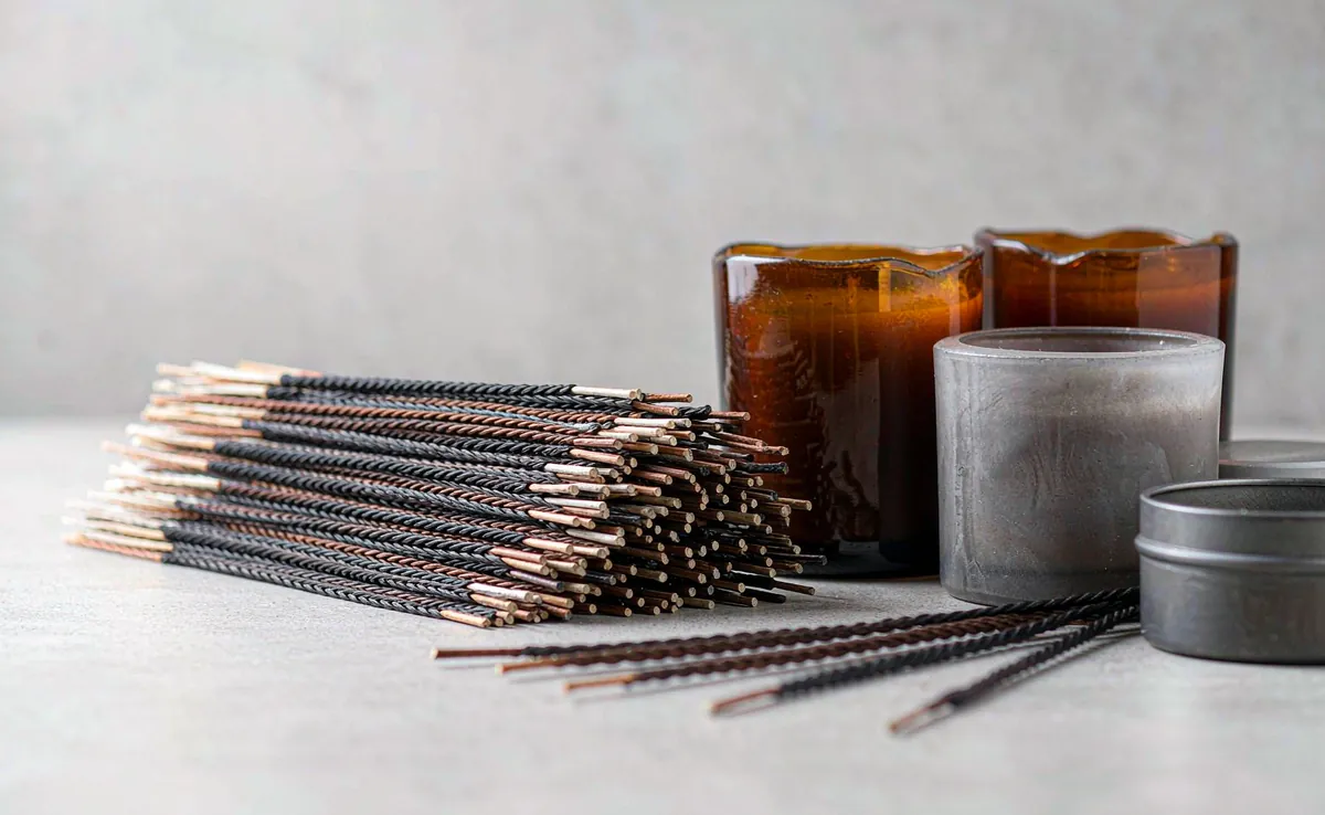 Cotton and wooden candle wicks beside amber glass jars, ceramic vessels and a tin container on an OEM scented candle production line